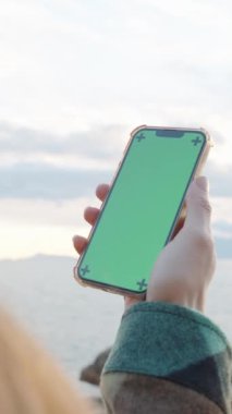 Smartphone with a Green Screen Mockup is holding by a Woman Against the Backbackground of the Sea and the Sun Behind Clouds.