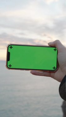 Bakış açısı. Yatay oryantasyon. Deniz kenarındaki uçuruma bakan bir kadının elinde boş yeşil ekranın maketi olan akıllı telefon. Güzel Manzaralar Yakalıyor.