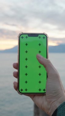 İlk-Kişi Görünümü, Bir Kadın Yeşil Ekranlı Akıllı Telefonu Tutar, Arıyor ve Parmaklarıyla Arıyor. Arka planda dağlar var. Denizin üzerinde bir gün batımı. Yüzüne yaklaştırıyor..