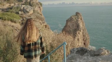 Bir kadın, deniz kenarındaki uçurum boyunca bilinmeyen yerleri keşfeder. Merdivenlerden aşağı iniyor..