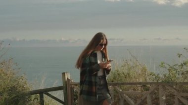 Genç, uzun saçlı bir kadın yüksek bir dağda deniz boyunca yürür, yoğun bir şekilde telefonuna bakar. Gün batımı.