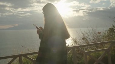 Deniz kıyısındaki bir uçurumun kenarında bir kadın, dağları ve arka plandaki suyun üzerindeki güneş ışınlarını gören bir kadın, telefonuna bakar..