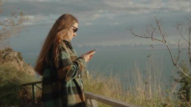 Kıyı Rüzgarı, Deniz kenarındaki bir uçurumun kenarında duran Genç Bir Kadının Uzun Saçlarını Tökezletir ve Akıllı Telefonuna bakar..