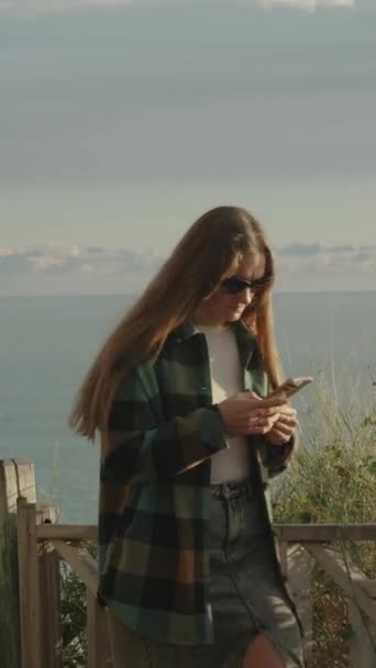 長髪の若い女性は 彼女の携帯電話を強く見て 高い山で海を歩いています サンセット — ストック動画
