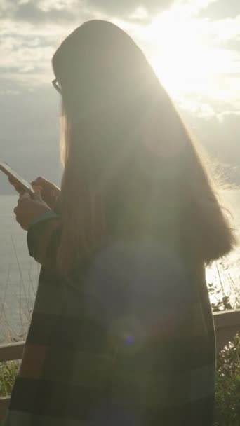 海の崖の端の女性 背景の水の上の山と太陽のレイと 彼女の携帯を見る — ストック動画