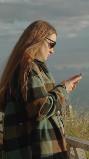 海岸の風は 海の崖の端に立っている若い女性の長い髪を痛めつけ 彼女のスマートフォンを見て — ストック動画