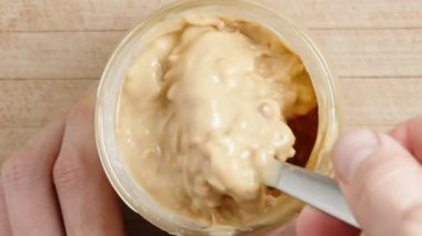 Kavanozda fıstık ezmesini kaşıkla karıştırmak, ahşap bir masanın üst manzarası. Yakın plan..