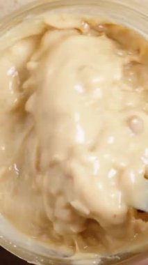 Dikey video. Kavanozda fıstık ezmesini kaşıkla karıştırmak, ahşap bir masanın üst manzarası. Yakın plan..