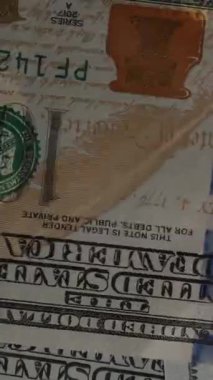 Dikey video. Kamera aşağıdan, cam masanın altından yüz dolarlık banknotlar topluyor. Mavi bir arkaplanda.