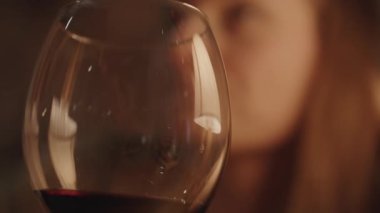 Bir bardak kırmızı şaraptaki masa lambasının yansıması, Genç bir kadın bir yudum alıp kameraya bir şey söylüyor..