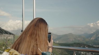 Uzun saçlı bir kadın akıllı telefonuyla dağları yakalıyor. Yüksek bir tepede, açık havada dikiliyor..