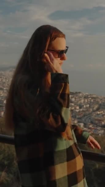 バーティカルビデオ チェッカーのシャツを着た若い女性が 夕暮れの間 街と海を背景に山の上を歩いている — ストック動画