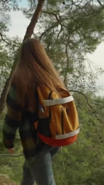 バーティカルビデオ 山の森の高い道に沿って彼女のショルダーウォークにバックパックを持つヤングトラベラー シティと海のエレベータービューを賞賛 — ストック動画