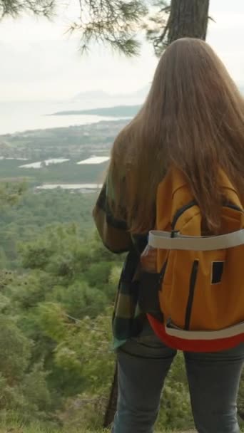 バーティカルビデオ 若い女性は 森を通って高い山に向かう道路の崖の端に立って 彼女のスマートフォンの距離で都市と海の景色をキャプチャ — ストック動画