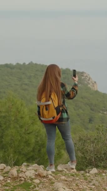 バーティカルビデオ 長髪の若い女性が首尾よく肩にバックパックで山頂に登った今 彼女のスマートフォンの海の美しい景色をキャプチャします — ストック動画