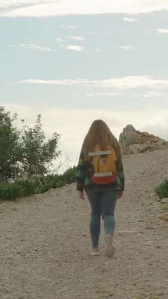 バーティカルビデオ 山頂への急な道に登る若い女性の後 古代遺跡は崖の近くにあった 日没は雲を通して暖かい光を投げます — ストック動画
