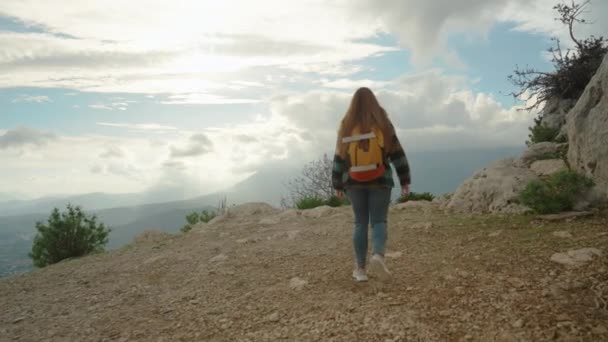 バックパックを持つ若い女性は 山頂の岩の上に登り 降雨の端に立って 山を見下ろす雲で映画の空と日没を楽しんでいます — ストック動画