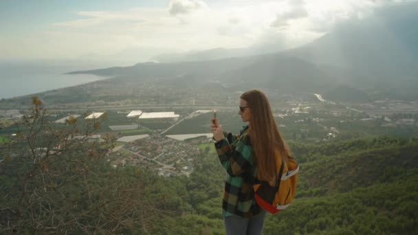 バックパックを持つ長髪の女性は世界を旅し 高い崖からスマートフォンで山と海の景色を捉え 深淵の端に立っています — ストック動画
