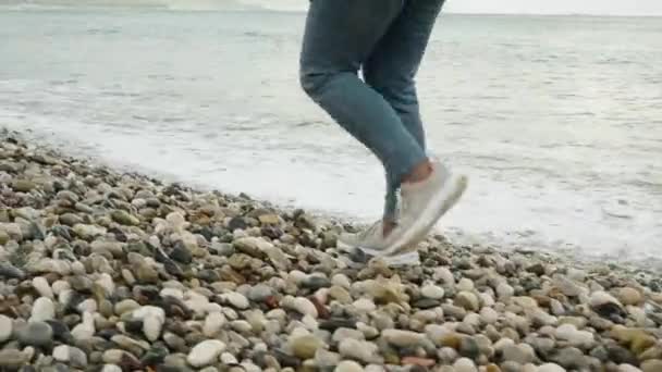 スニーカーとジーンズの女性はペブルビーチに沿って歩きます 波はほぼ彼女の足に触れる — ストック動画