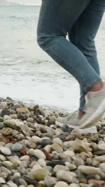 一个穿着短裤和牛仔裤的女人沿着卵石滩散步 波浪几乎触动了她的脚 — 图库视频影像