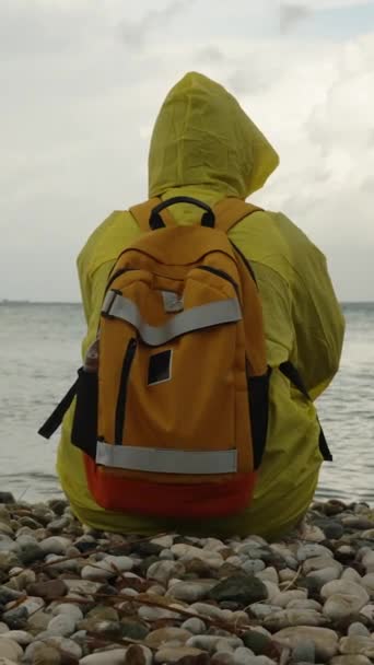 从后面看一个穿着黄色雨衣 背着橙色背包的人 坐在汹涌大海边的海滩上 旅行人员 — 图库视频影像