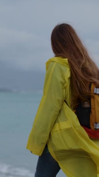 海上阴郁的天气 在寒冷的蓝调中发亮 穿着鲜艳的黄色雨衣背着橙色背包在海滩上散步的年轻女子 慢动作 — 图库视频影像