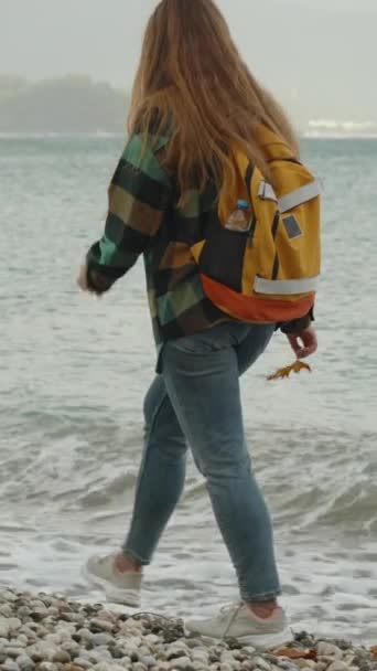 オレンジ色のバックパックを持った若い女性は 彼女の手でメープル秋の葉を保持し 天気のオーバーキャストでビーチを散歩 — ストック動画