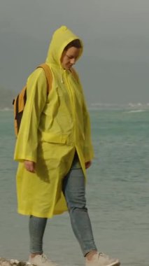 Bulutlu havada parlak sarı yağmurluk giyen genç bir kadın, deniz kenarında yavaşça yürür. Sırtında turuncu bir sırt çantası var..