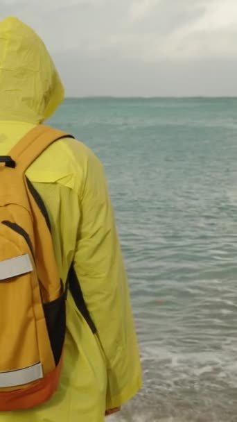 一个孤零零的人 穿着鲜艳的黄色雨衣 背着背包 站在那里 凝视着远方汹涌的大海和阴沉沉的天空 — 图库视频影像