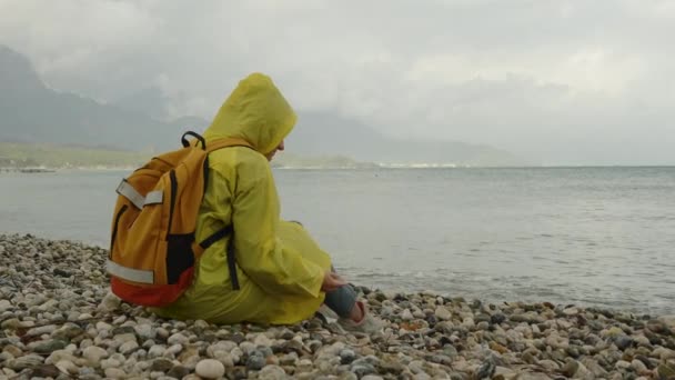 黄色いレインコートにバックパックが入った女性が座り 海に石を投げ込む 雨の天気と天空を覆っている 孤独と悲しみ — ストック動画