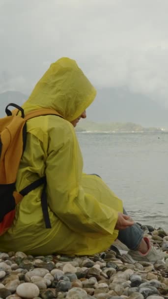 一个穿着黄色雨衣 背着背包的女人坐在那里 把一块石头扔进了海里 多雨的天气和阴暗的天空 孤独和悲伤 — 图库视频影像