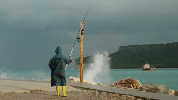 一个穿着蓝色雨衣和黄色靴子的渔夫手里拿着钓竿站在海上的风暴中 强烈的海浪冲击着附近的岩石 在远方 一艘船和群山在海湾 — 图库视频影像