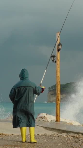 一个穿着蓝色雨衣和黄色靴子的渔夫手里拿着钓竿站在海上的风暴中 强烈的海浪冲击着附近的岩石 在远方 一艘船和群山在海湾 — 图库视频影像