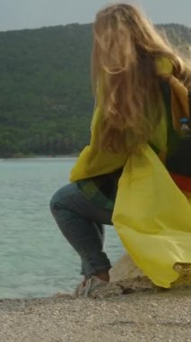 Sarı ceketli genç bir kadın deniz kenarındaki bir kayaya oturur, fırtınalı hava kıyafetlerini ve saçlarını dalgalandırırken, güneş ışınları bulutları delip geçerken, onu arkadan aydınlatır..