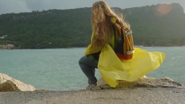 一位身穿黄色外套的年轻女子坐在海边的岩石上 暴风雨般的天气吹拂着她的衣服和头发 阳光穿透了云层 从后面照射着她 — 图库视频影像
