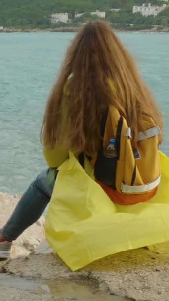 在恶劣的天气里 一个穿着黄色雨衣 背着橙色背包的女人坐在汹涌大海边的岩石上 蓝色的水 — 图库视频影像