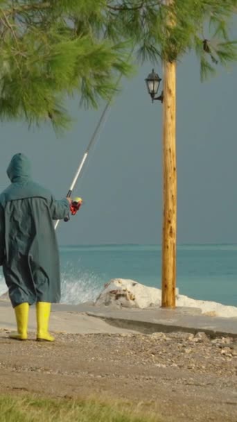 一个渔夫穿着雨衣和黄色的靴子 在暴风雨中的海滨长廊上钓鱼 大浪在附近的岩石上猛烈冲撞 远山高耸 — 图库视频影像