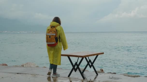 一个穿着黄色雨衣的女人在海滨码头上的暴风雨天气里 走近一家关着的咖啡店的桌边 — 图库视频影像