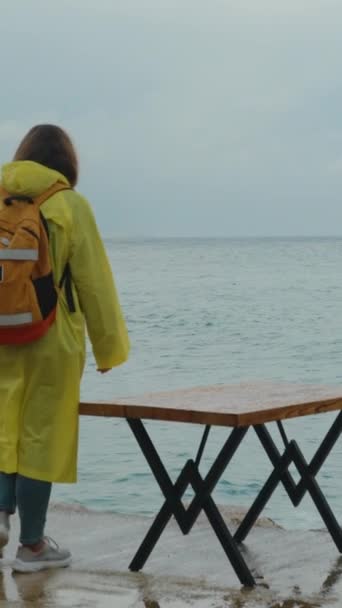 海岸の桟橋で嵐の天候で黄色いレインコートの女性は 閉じたカフェのテーブルの近くの端に近づきます — ストック動画