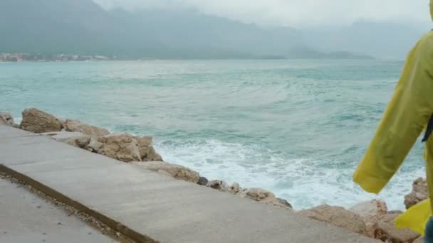 一个穿着黄色雨衣的女人带着头巾和背包 在暴风雨中沿着码头走着 大浪正冲向她 — 图库视频影像