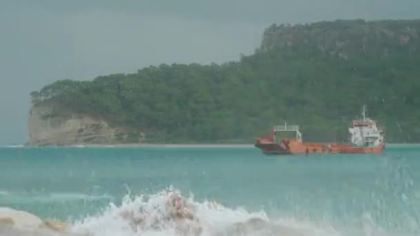 一艘货船在海湾的波浪上摇晃着 蓝色的海浪冲击着岩石 产生了微小的水花 背景上有茂密森林的悬崖 — 图库视频影像