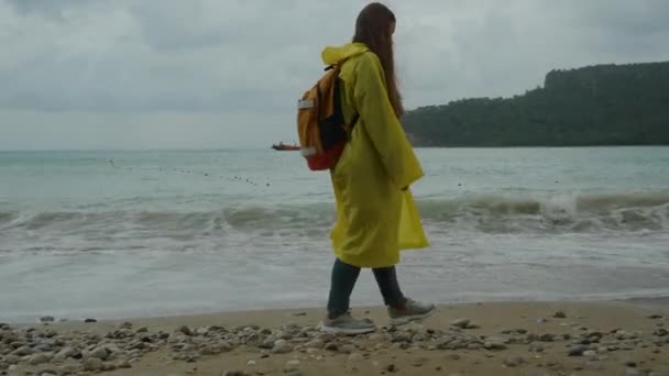 一个背着背包的女人穿着一件黄色雨衣在海滩上散步 — 图库视频影像