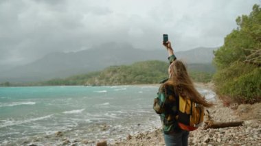 Deniz kenarındaki sert rüzgâr, akıllı telefonuyla fotoğraf çekerken sırt çantalı genç bir seyahat blogcusunun uzun saçlarını okşuyor. Yavaş çekim.