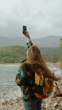 Dikey video. Deniz kenarındaki sert rüzgâr, akıllı telefonuyla fotoğraf çekerken sırt çantalı genç bir seyahat blogcusunun uzun saçlarını okşuyor. Yavaş çekim.