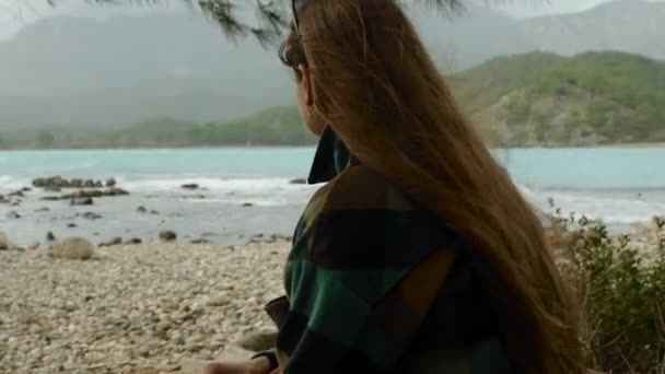 在寒冷的天气里 一个女人独自呆在一个空旷的海滨 躲藏在松树枝下 在高山的背景下欣赏海湾里碧绿的水 女权主义者的概念 在荒芜的海滩上自由自在 — 图库视频影像