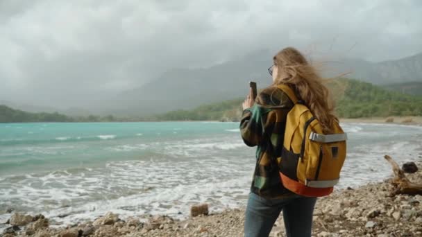 ターコイズの海水と山の背景に対する波 若い女性のリアビュー 旅行ブロガー ソーシャルメディアの写真を撮る または保管庫として 強い風は彼女の長い髪を悩ませました — ストック動画