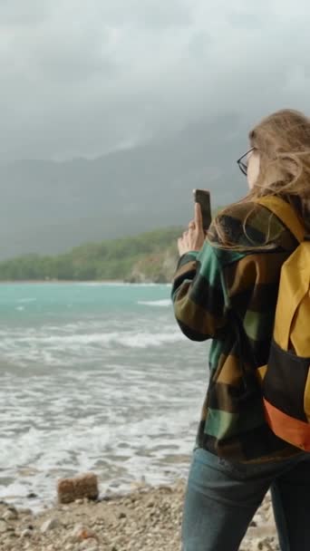 バーティカルビデオ ターコイズの海水と山の背景に対する波 若い女性のリアビュー 旅行ブロガー ソーシャルメディアの写真を撮る または保管庫として 強い風は彼女の長い髪を悩ませました — ストック動画