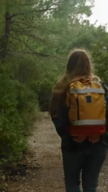 Dikey video. Turuncu bir sırt çantasıyla yürüyüş yapan ve ormanda tek başına gezen genç bir kadının arka görüntüsü. Hava bulutlu ve serin.