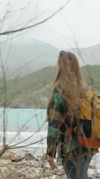 バーティカルビデオ バックパック付きの女性観光客は 寒い秋の日にターコイズ海水と山々で岩場沿いに散歩します スローモーション — ストック動画