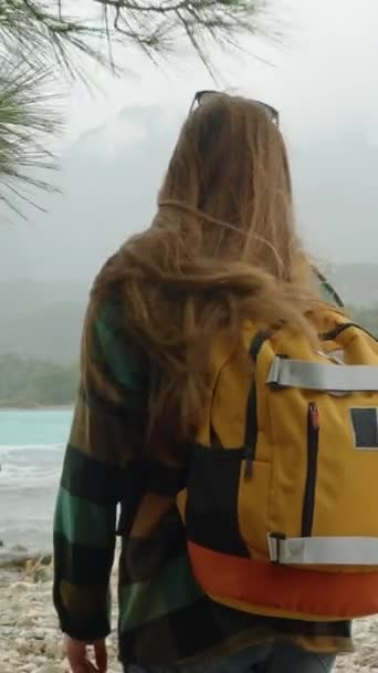 バーティカルビデオ 海の湾に沿って松林を巡って 寒い日を過ごす オレンジ色のバックパックが歩いている女性 — ストック動画